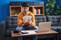 Young hispanic man using laptop holding dollars sitting on sofa at home Royalty Free Stock Photo