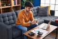 Young hispanic man using laptop holding dollars sitting on sofa at home Royalty Free Stock Photo