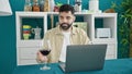 Young hispanic man using laptop holding glass of wine at dinning room Royalty Free Stock Photo