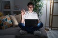 Young hispanic man using laptop at home at night waiving saying hello happy and smiling, friendly welcome gesture Royalty Free Stock Photo