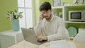 Young hispanic man using laptop talking on smartphone sitting on table at dinning room Royalty Free Stock Photo