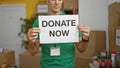 Young hispanic man volunteer holding donate now banner at charity center