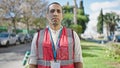 Young hispanic man volunteer wearing vest looking serious at park