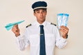 Young hispanic man wearing airplane pilot uniform holding paper airplane and boarding pass in shock face, looking skeptical and Royalty Free Stock Photo