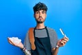 Young hispanic man wearing barber apron holding razor and foam puffing cheeks with funny face