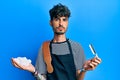 Young hispanic man wearing barber apron holding razor and foam relaxed with serious expression on face