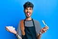 Young hispanic man wearing barber apron holding razor and foam sticking tongue out happy with funny expression