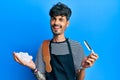 Young hispanic man wearing barber apron holding razor and foam winking looking at the camera with sexy expression, cheerful and