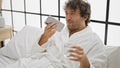 Young hispanic man wearing bathrobe drinking water sending voice message by smartphone at bedroom