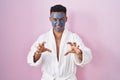 Young hispanic man wearing beauty face mask and bath robe smiling funny doing claw gesture as cat, aggressive and sexy expression Royalty Free Stock Photo