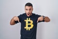 Young hispanic man wearing bitcoin t shirt pointing down with fingers showing advertisement, surprised face and open mouth