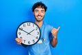 Young hispanic man wearing blue male nurse uniform holding clock pointing thumb up to the side smiling happy with open mouth Royalty Free Stock Photo