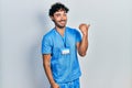 Young hispanic man wearing blue male nurse uniform smiling with happy face looking and pointing to the side with thumb up Royalty Free Stock Photo