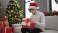 Young hispanic man wearing christmas hat unpacking gift with surprised face at home Royalty Free Stock Photo