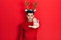 Young hispanic man wearing cute christmas reindeer horns doing stop sing with palm of the hand