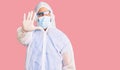 Young hispanic man wearing doctor protection coronavirus uniform and medical mask doing stop sing with palm of the hand Royalty Free Stock Photo