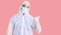 Young hispanic man wearing doctor protection coronavirus uniform and medical mask smiling with happy face looking and pointing to Royalty Free Stock Photo