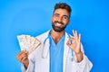 Young hispanic man wearing doctor uniform holding uk pounds banknotes doing ok sign with fingers, smiling friendly gesturing