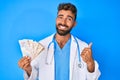 Young hispanic man wearing doctor uniform holding uk pounds banknotes smiling happy and positive, thumb up doing excellent and