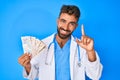 Young hispanic man wearing doctor uniform holding uk pounds banknotes smiling with an idea or question pointing finger with happy