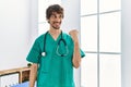 Young hispanic man wearing doctor uniform and stethoscope at clinic smiling with happy face looking and pointing to the side with Royalty Free Stock Photo