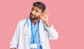 Young hispanic man wearing doctor uniform and stethoscope smiling with hand over ear listening an hearing to rumor or gossip Royalty Free Stock Photo