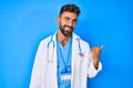 Young hispanic man wearing doctor uniform and stethoscope smiling with happy face looking and pointing to the side with thumb up Royalty Free Stock Photo