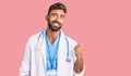 Young hispanic man wearing doctor uniform and stethoscope smiling with happy face looking and pointing to the side with thumb up Royalty Free Stock Photo