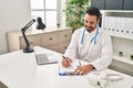 Young hispanic man wearing doctor uniform writing on clipboard at clinic Royalty Free Stock Photo