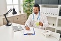Young hispanic man wearing doctor uniform writing on clipboard drinking coffee at clinic Royalty Free Stock Photo