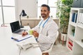 Young hispanic man wearing doctor uniform writing on clipboard drinking coffee at clinic Royalty Free Stock Photo