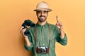 Young hispanic man wearing explorer hat looking through binoculars smiling with an idea or question pointing finger with happy Royalty Free Stock Photo