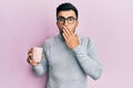 Young hispanic man wearing glasses and drinking a cup of coffee covering mouth with hand, shocked and afraid for mistake Royalty Free Stock Photo