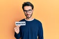 Young hispanic man wearing glasses holding winter word on paper thinking attitude and sober expression looking self confident
