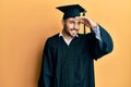 Young hispanic man wearing graduation cap and ceremony robe very happy and smiling looking far away with hand over head Royalty Free Stock Photo