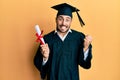 Young hispanic man wearing graduation robe holding diploma screaming proud, celebrating victory and success very excited with Royalty Free Stock Photo