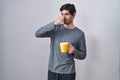 Young hispanic man wearing pajama drinking a cup of coffee smelling something stinky and disgusting, intolerable smell, holding