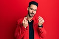 Young hispanic man wearing red leather jacket doing money gesture with hands, asking for salary payment, millionaire business