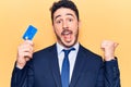 Young hispanic man wearing suit holding credit card pointing thumb up to the side smiling happy with open mouth Royalty Free Stock Photo