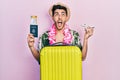 Young hispanic man wearing summer style and hawaiian lei holding passport and plane toy angry and mad screaming frustrated and Royalty Free Stock Photo