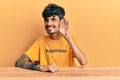 Young hispanic man wearing tshirt with happiness word message sitting on the table smiling with hand over ear listening an hearing