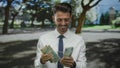Young hispanic man in white shirt counting dirham banknotes with hands in forest clearing; prosperity Royalty Free Stock Photo