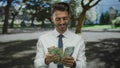 Young hispanic man in white shirt counting dirham banknotes with hands in forest clearing; prosperity Royalty Free Stock Photo