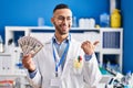 Young hispanic man working at scientist laboratory holding money pointing thumb up to the side smiling happy with open mouth Royalty Free Stock Photo