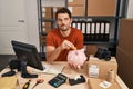 Young hispanic man working at small business ecommerce holding piggy bank smiling looking to the side and staring away thinking Royalty Free Stock Photo