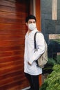 Young Hispanic medical student wearing a facemask and a medical robe and leaving his house