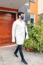 Young Hispanic medical student wearing a facemask and a medical robe while leaving the hospital