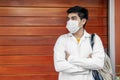 Young Hispanic medical student wearing a medical robe and a facemask while leaning on a wooden wall