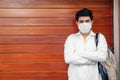 Young Hispanic medical student wearing a medical robe and a facemask while leaning on a wooden wall