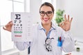 Young hispanic optician woman holding medical exam doing ok sign with fingers, smiling friendly gesturing excellent symbol Royalty Free Stock Photo